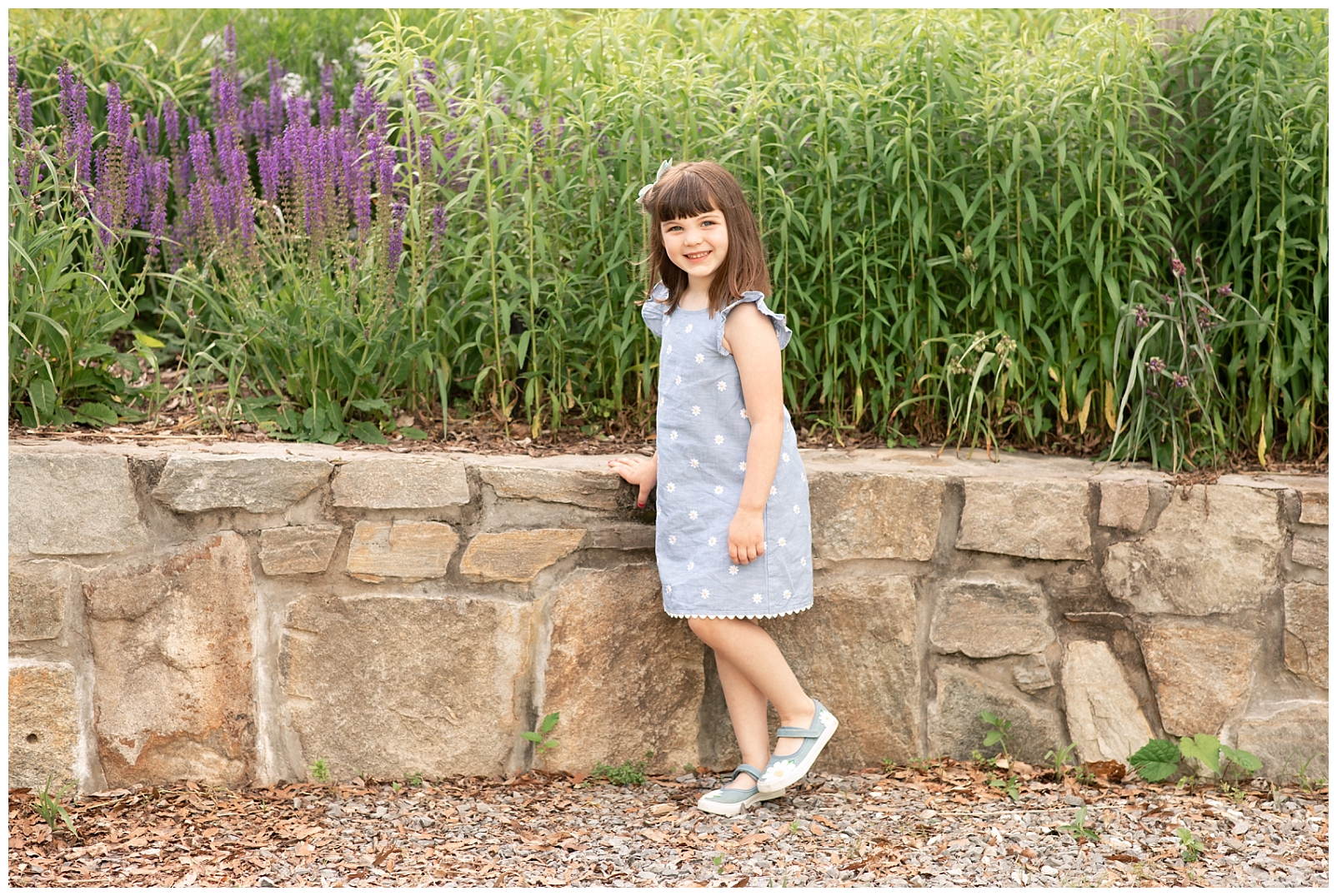 girl standing near a stone wall with purple flowers