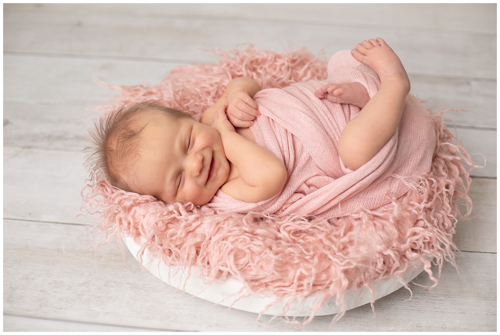 Newborn girl in pink wrap on a pink fluffy faux fur fabric on a wooden bowl