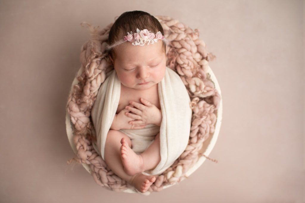 Newborn wrapped in a cream wrap on a mauve stitched background in a round bowl with a floral headband