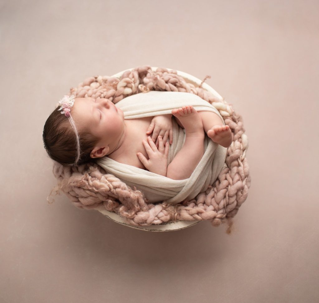 Newborn wrapped in a cream wrap on a mauve stitched background in a round bowl with a floral headband