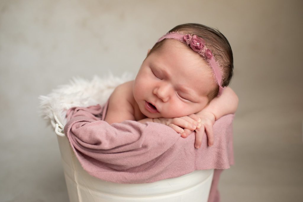 Newborn with chin on hands in bucket with a mauve floral headband