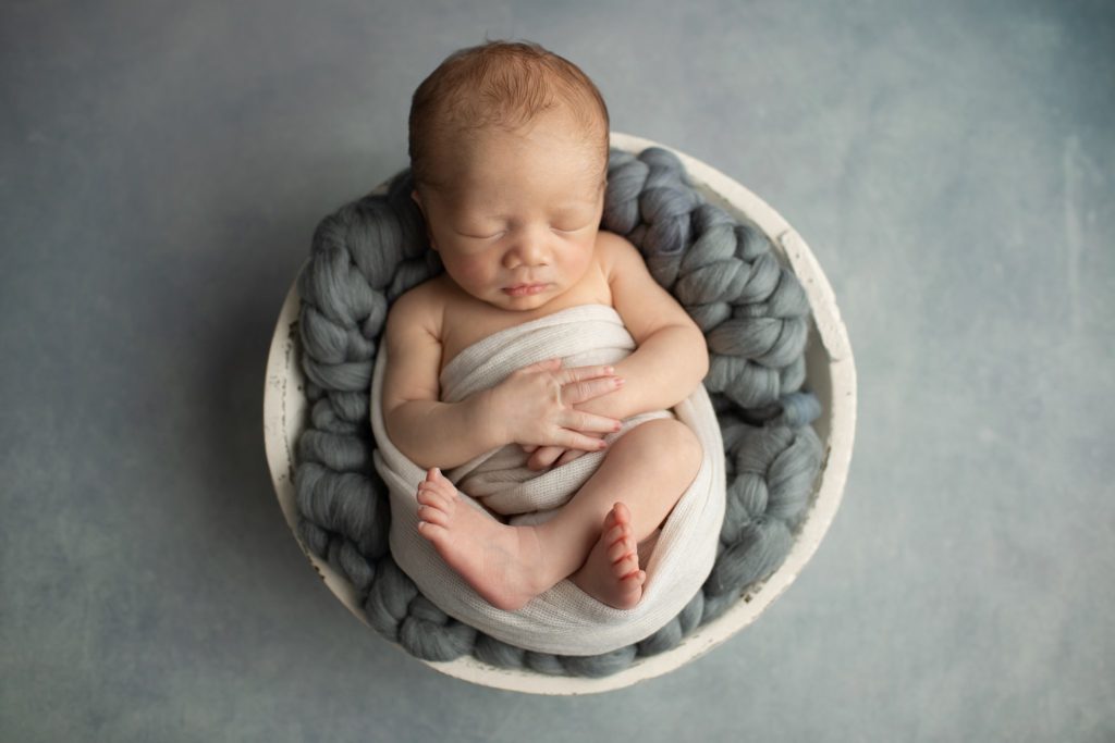 Newborn boy in a bowl with chunky gray wool knit blanket