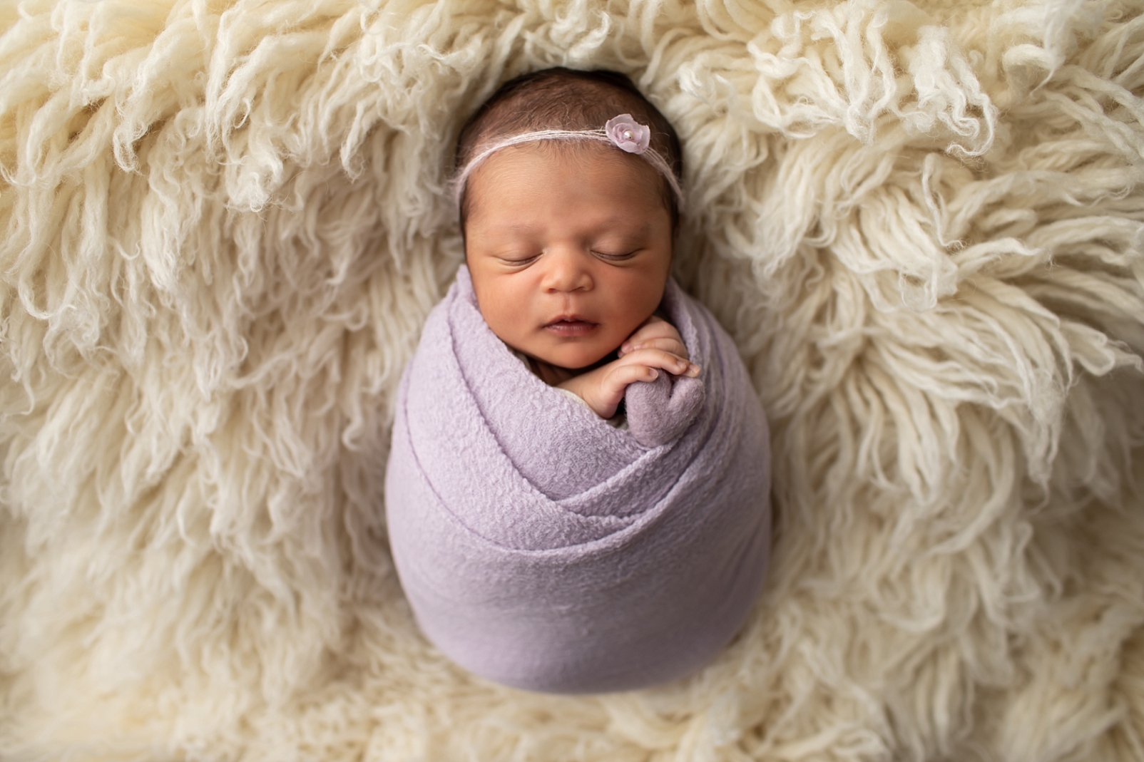 Newborn girl wrapped in lavender wrap and holding a heart on flokati