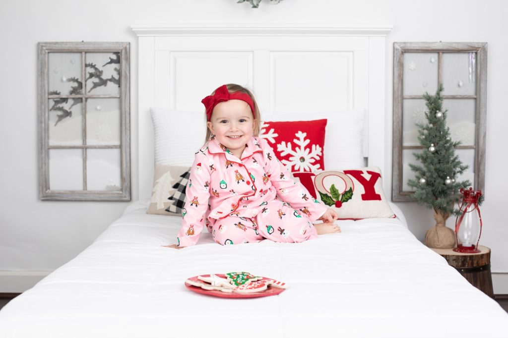 Little girl in pink flannel pajamas with Christmas pattern sitting on a bed