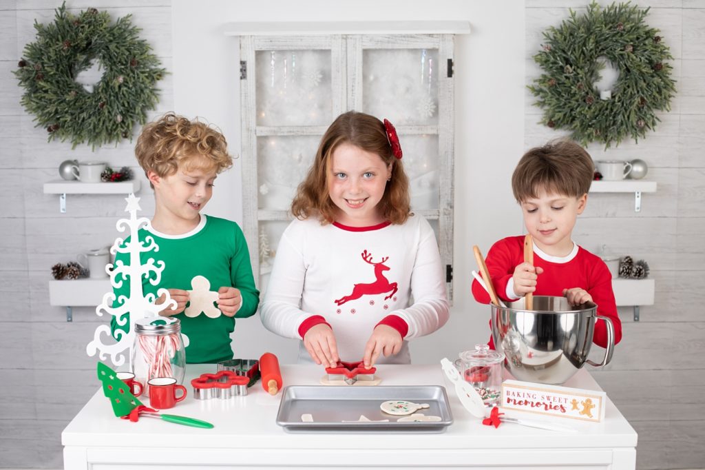 Three children making salt dough Christmas cookies in a room with shelves and a window with snow outside
