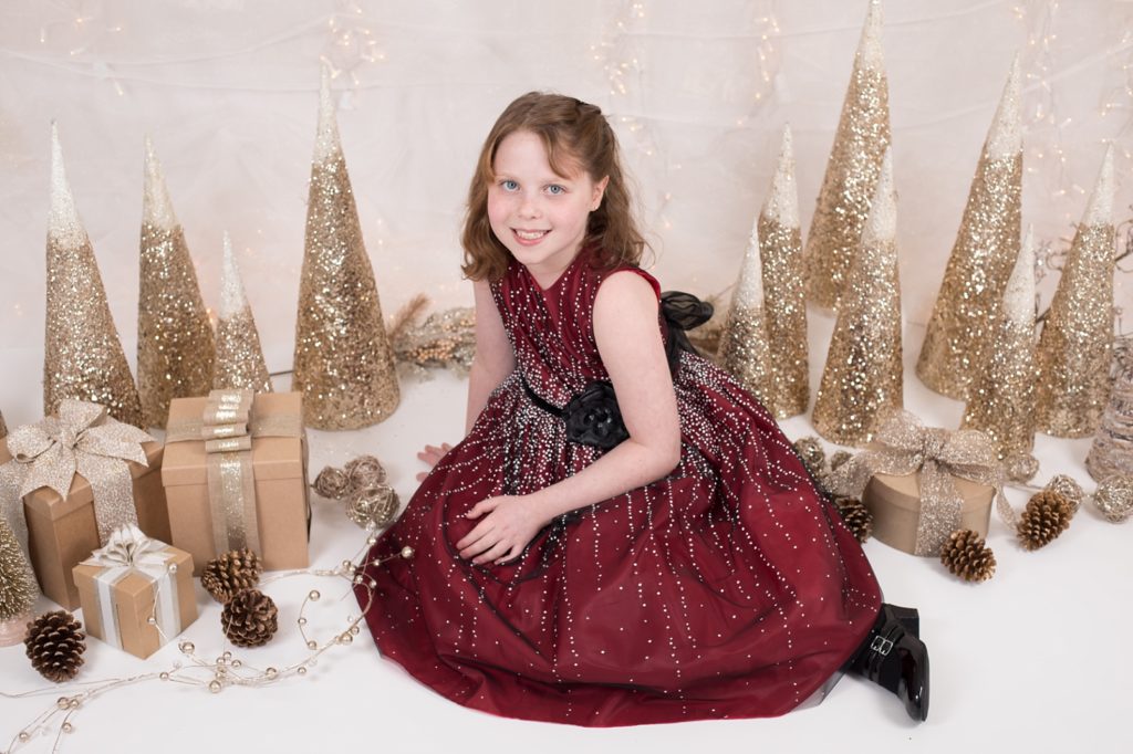 girl sitting in burgundy color dress in front of gold glitter cone trees and brown kraft paper gift boxes adorned with beautiful ribbons
