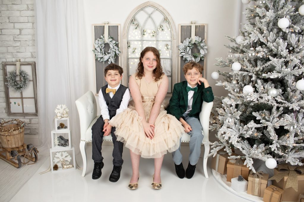 Three children sitting on a bench in front of a window and next to a Christmas tree
