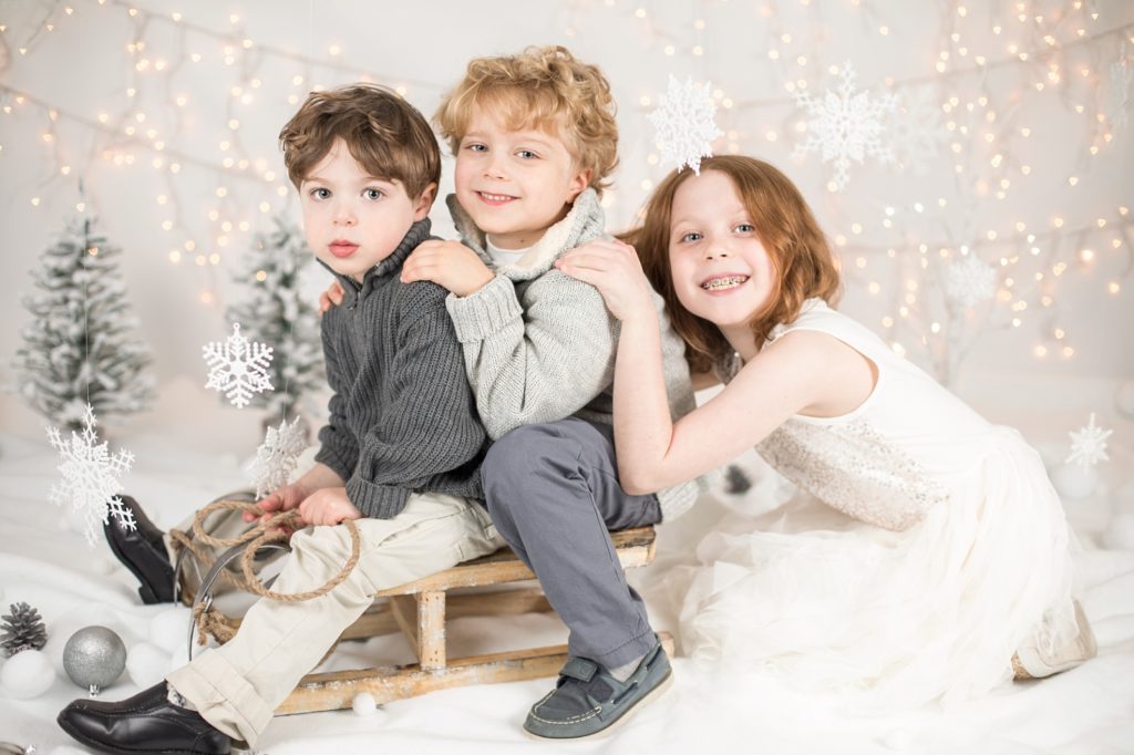 Two boys sitting on a little sled with snowflakes falling from the ceiling and bokeh Christmas lights in the background. And a girl in a white tulle gown smiling
