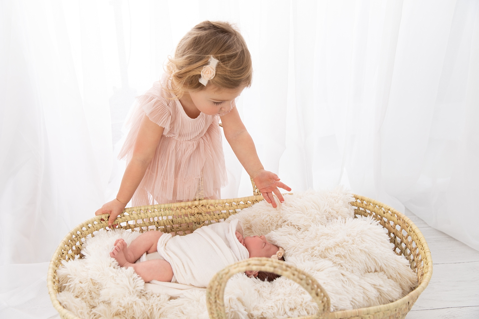 girl in pink dress in front of white curtains looking at newborn sibling in a basket