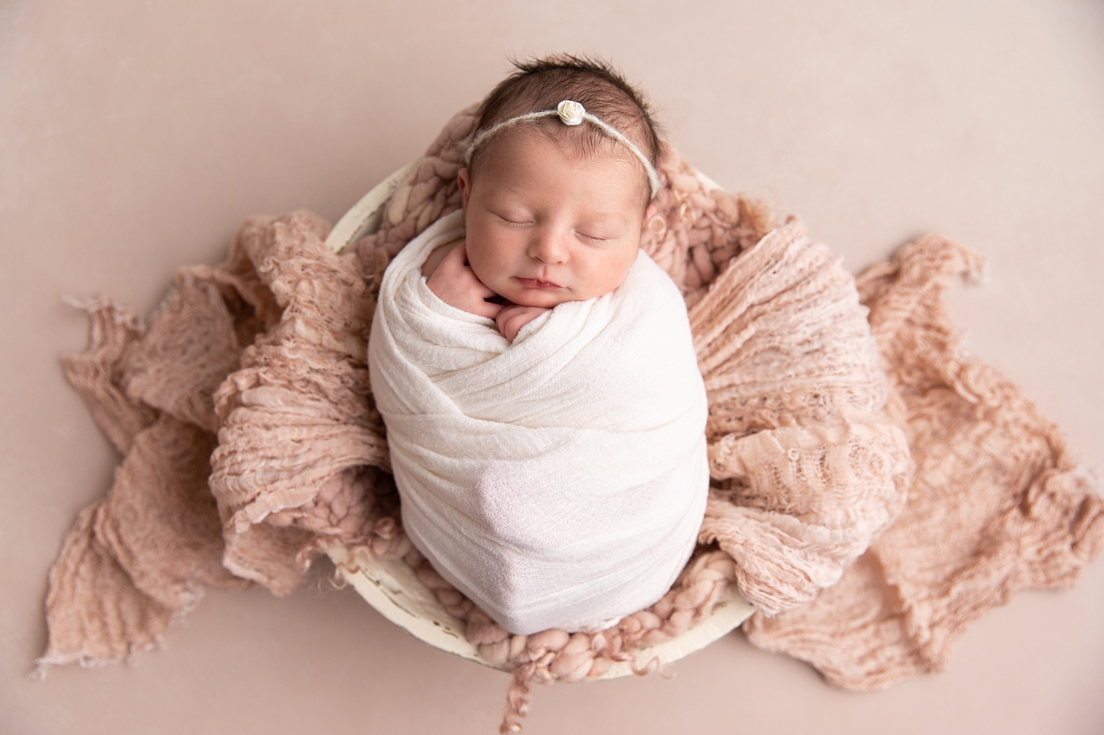 Newborn girl swaddled with decorative headband and lying in a blush fabric bowl