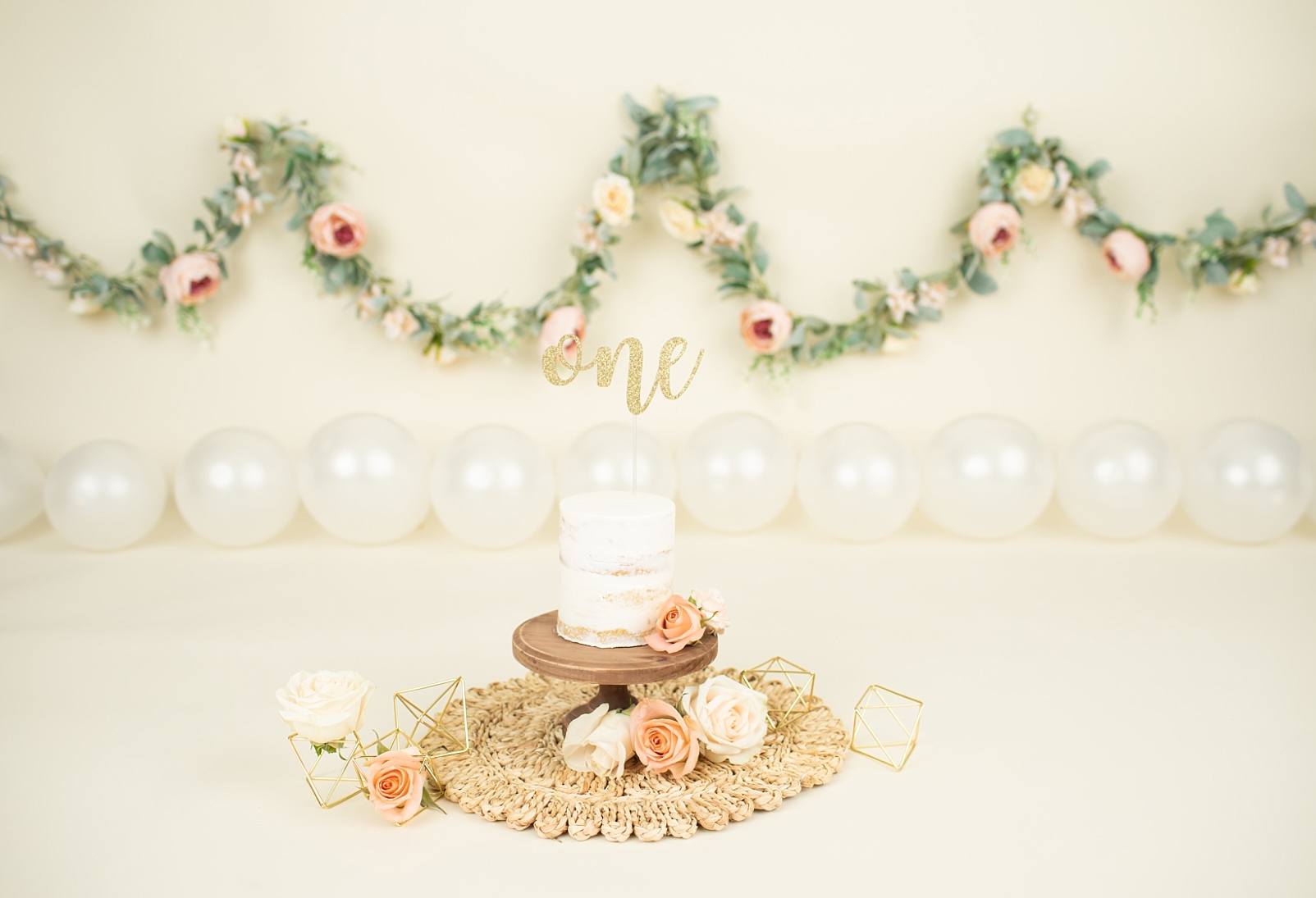 Cake smash cake with fresh flower adornment and gold geometric shapes and a floral garland background
