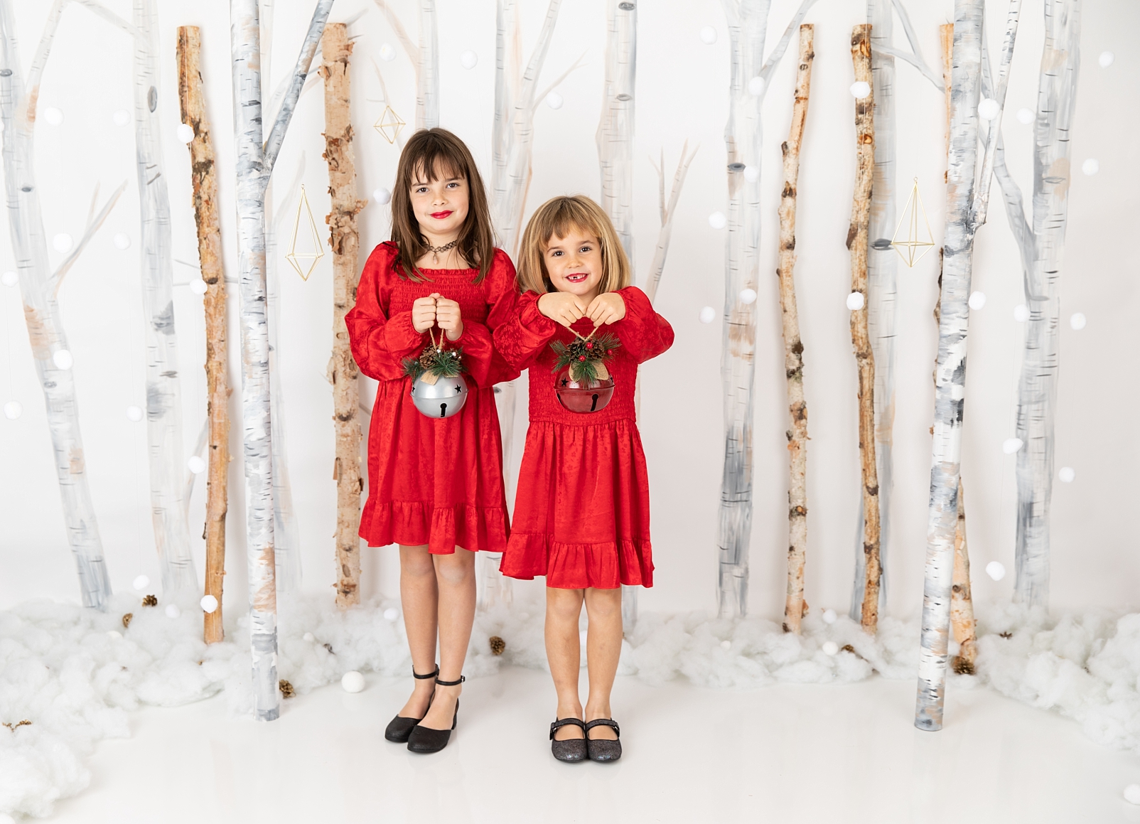 two girls in red dresses holding jingle bells in front of birch tree background
