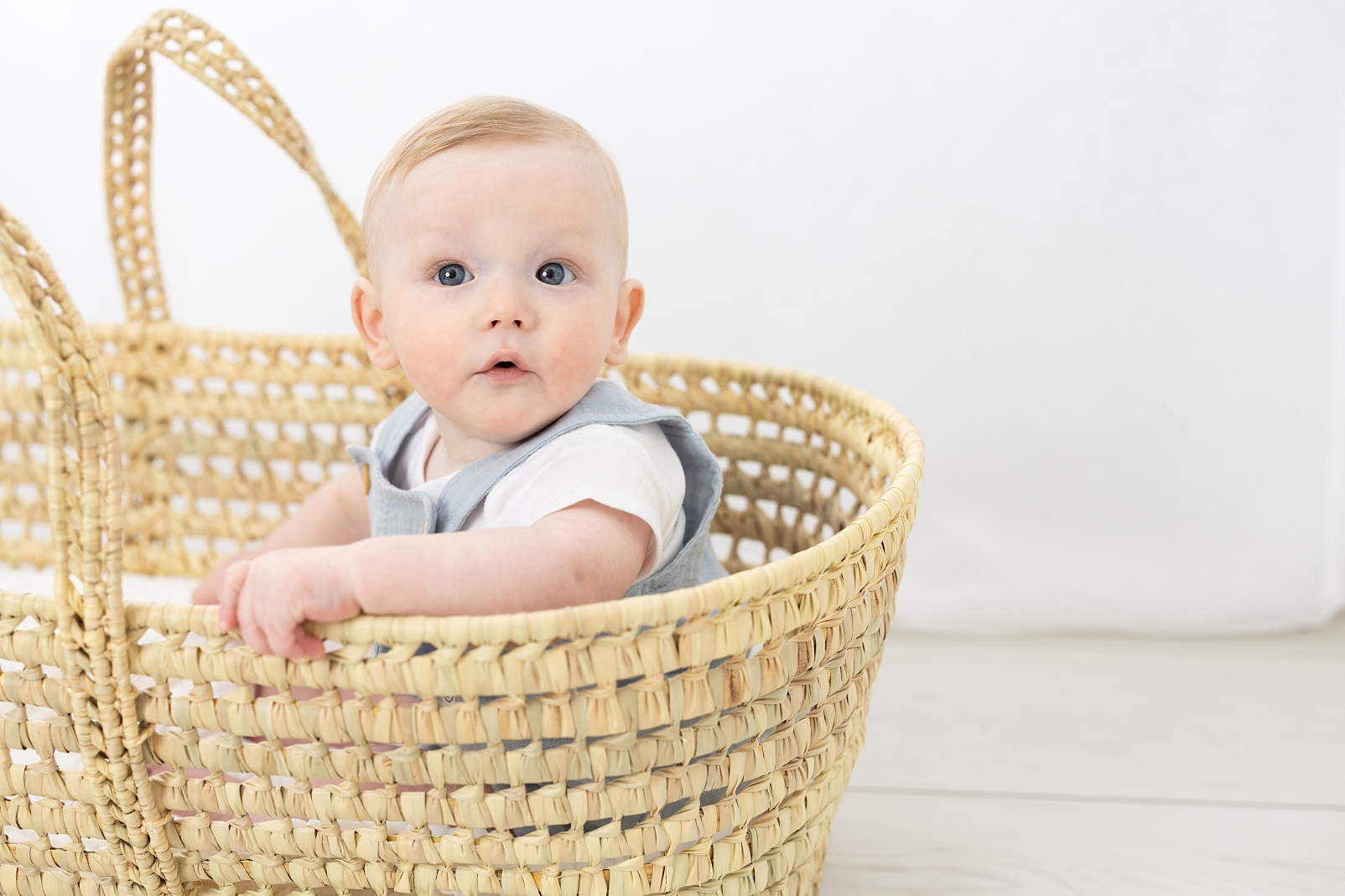 7 months milestone portraits of baby boy in light blue overalls sitting in a basket

