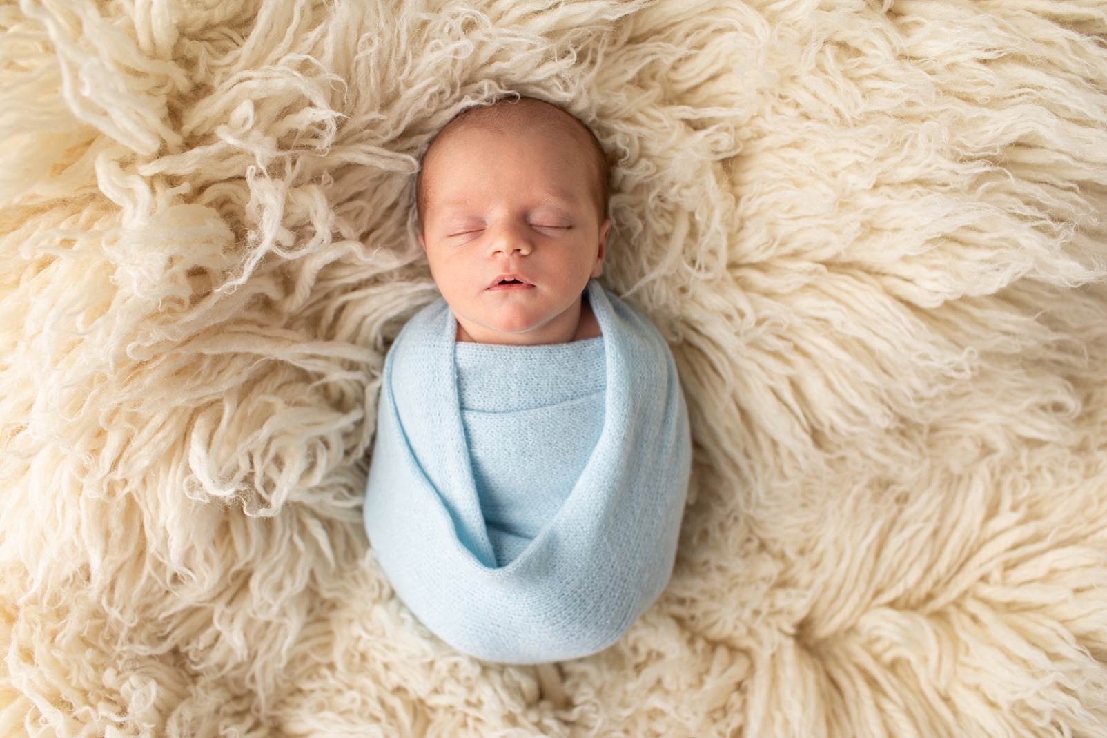 newborn baby boy wrapped in light blue wrap and sleeping on a flokati