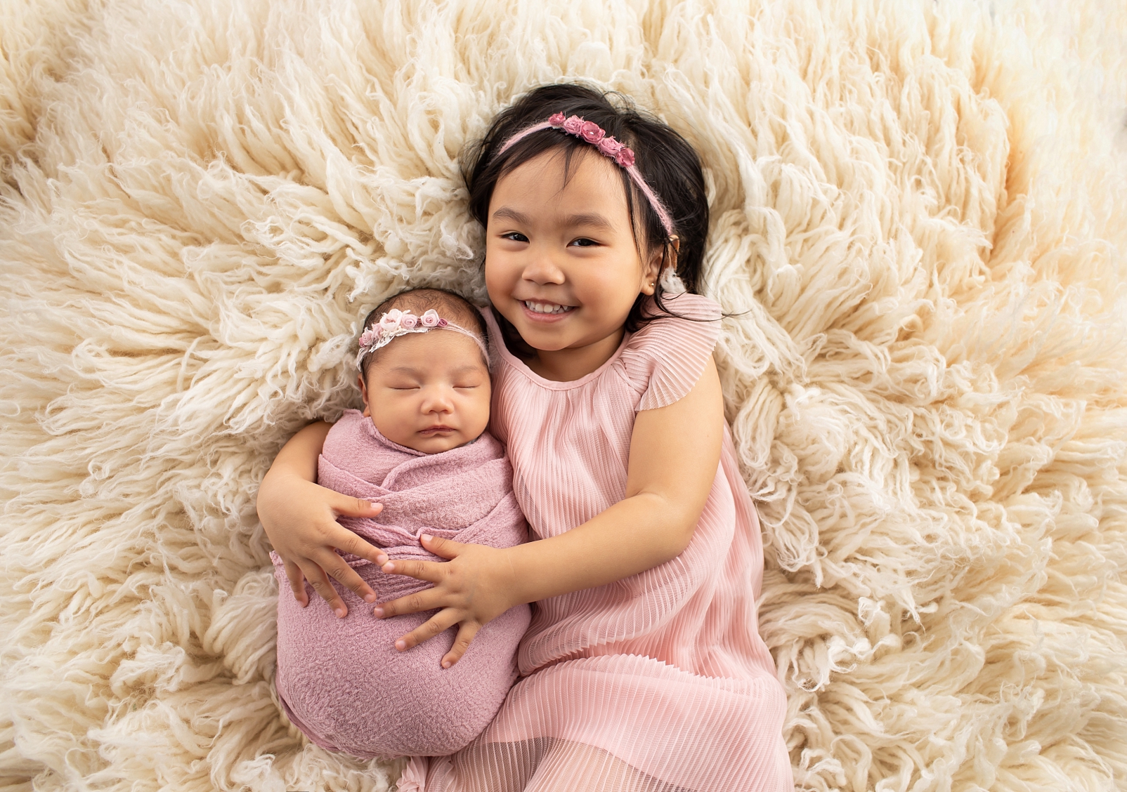 A newborn girl in a mauve color wrap with floral headband being hugged by her sister in a pink dress on a flokati rug for newborn portraits of a First Year Membership
