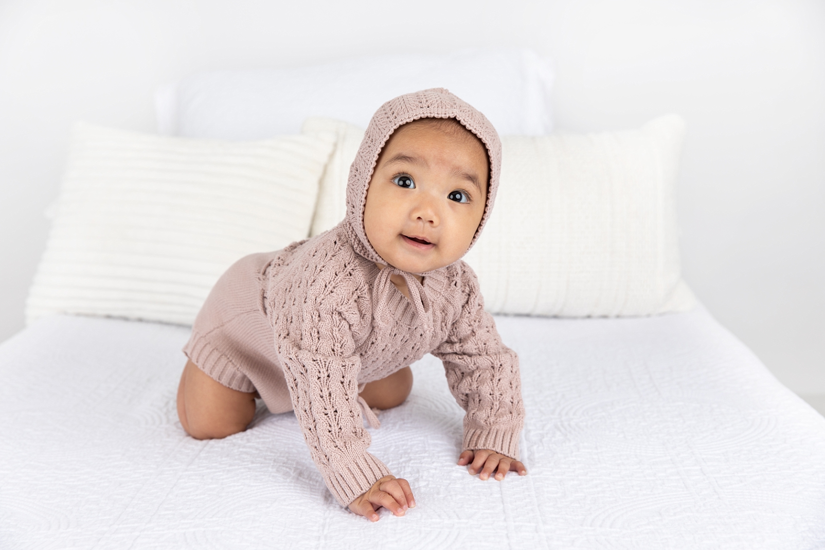 Baby girl in knit mauve outfit and bonnet smiling and crawling on white fabric as part of a First Year Membership | Maryland Baby Photographer