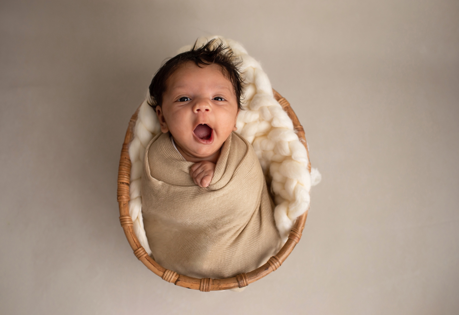 Baby yawns while happily looking at camera in a swaddle in a a basket