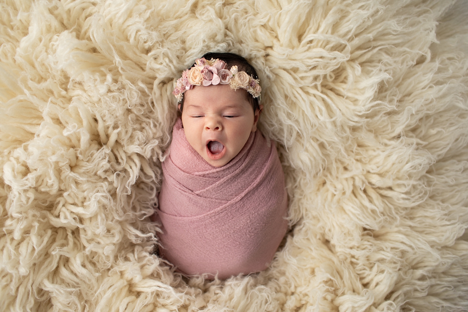 Baby yawns in a mauve wrap and floral headband on a flokati rug