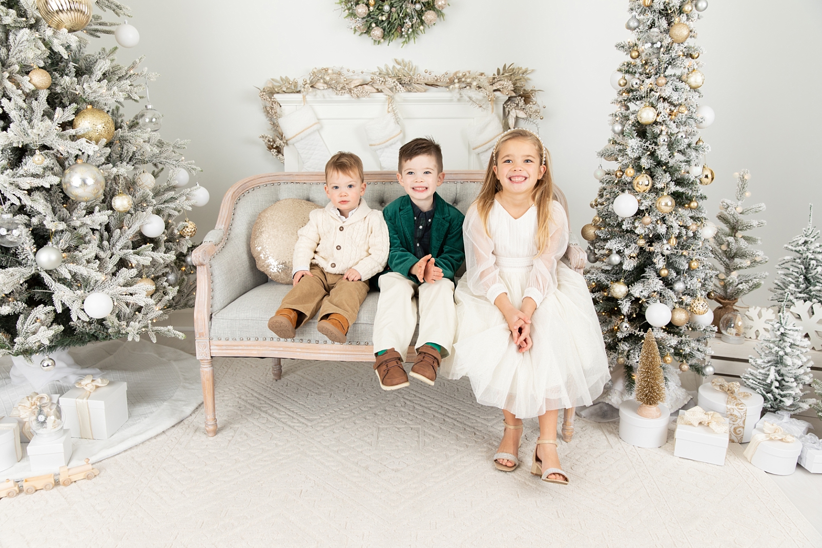 Three children sitting on a gray loveseat with a gold pillow in front of a fireplace mantle and stockings with decorated trees on either side for Christmas Sessions Maryland 2024