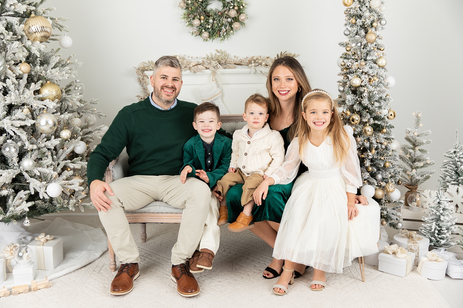 Family of 5 wearing green, neutrals, and white, sitting on loveseat in front of a fireplace mantle surrounded by silver, white, and gold decorated flocked Christmas trees for Christmas Sessions Maryland 2024 with Rebecca Leigh Photography