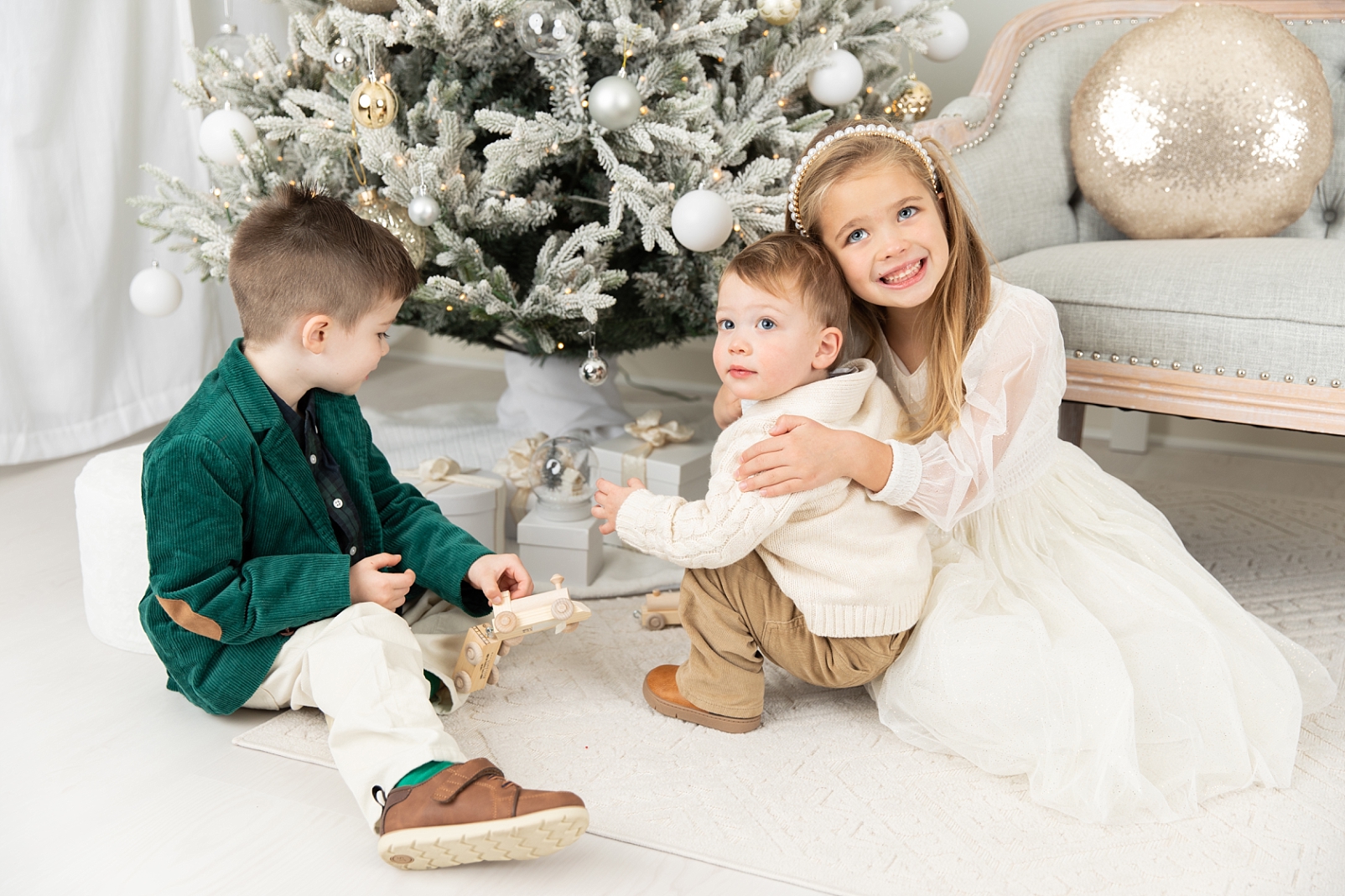 A girl and her 2 brothers playing with wooden trains in front of the Christmas tree
