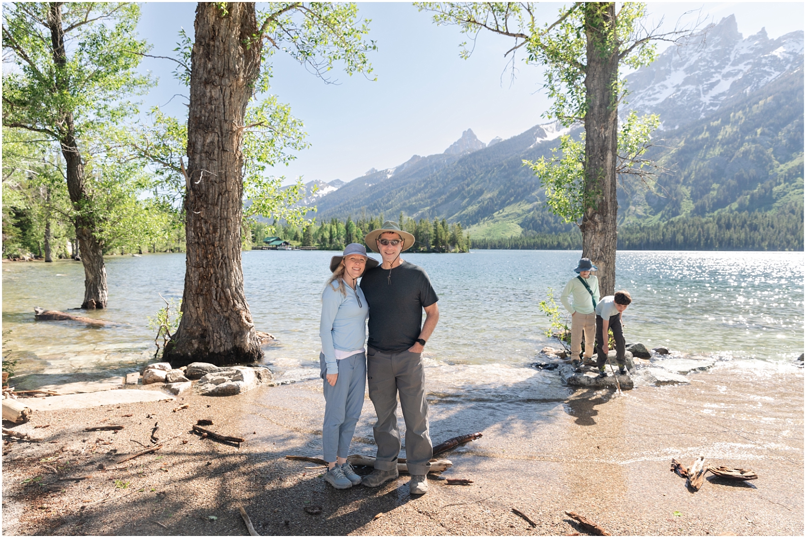 A couple standing in front of Jenny Lake with two boys playing in the background by the water - 3 Tips for Taking Your Own Family Photos by Rebecca Leigh Photography