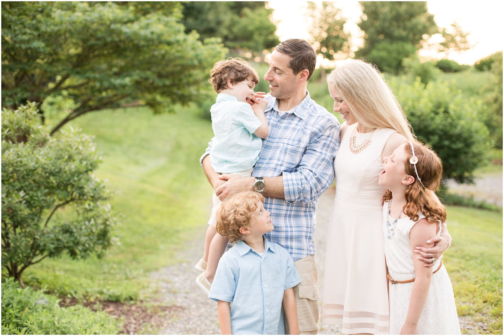 A mom and dad smiling at their 3 children, all wearing pastel colors, standing on a rocky pathway with green trees in the background - to demonstrate how to take your own family photos by Rebecca Leigh Photography