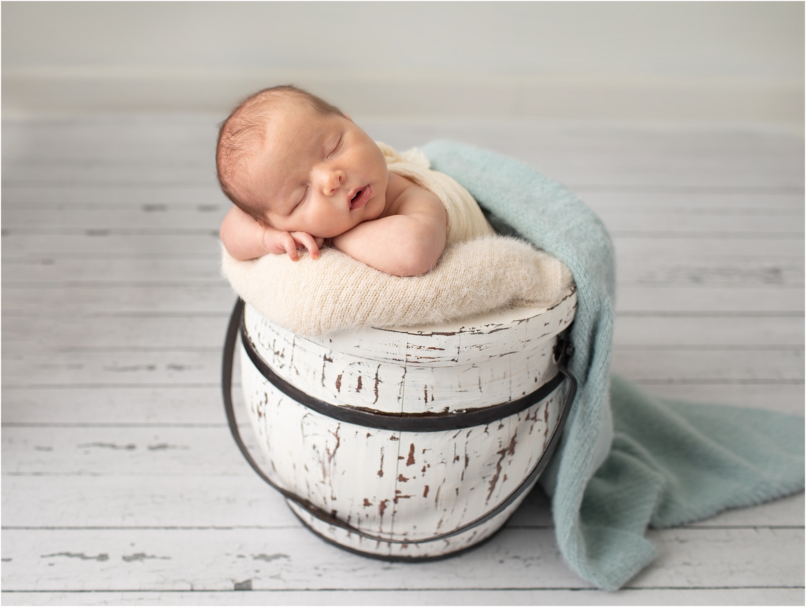 Newborn baby boy sleeping with face on arms in white bucket.