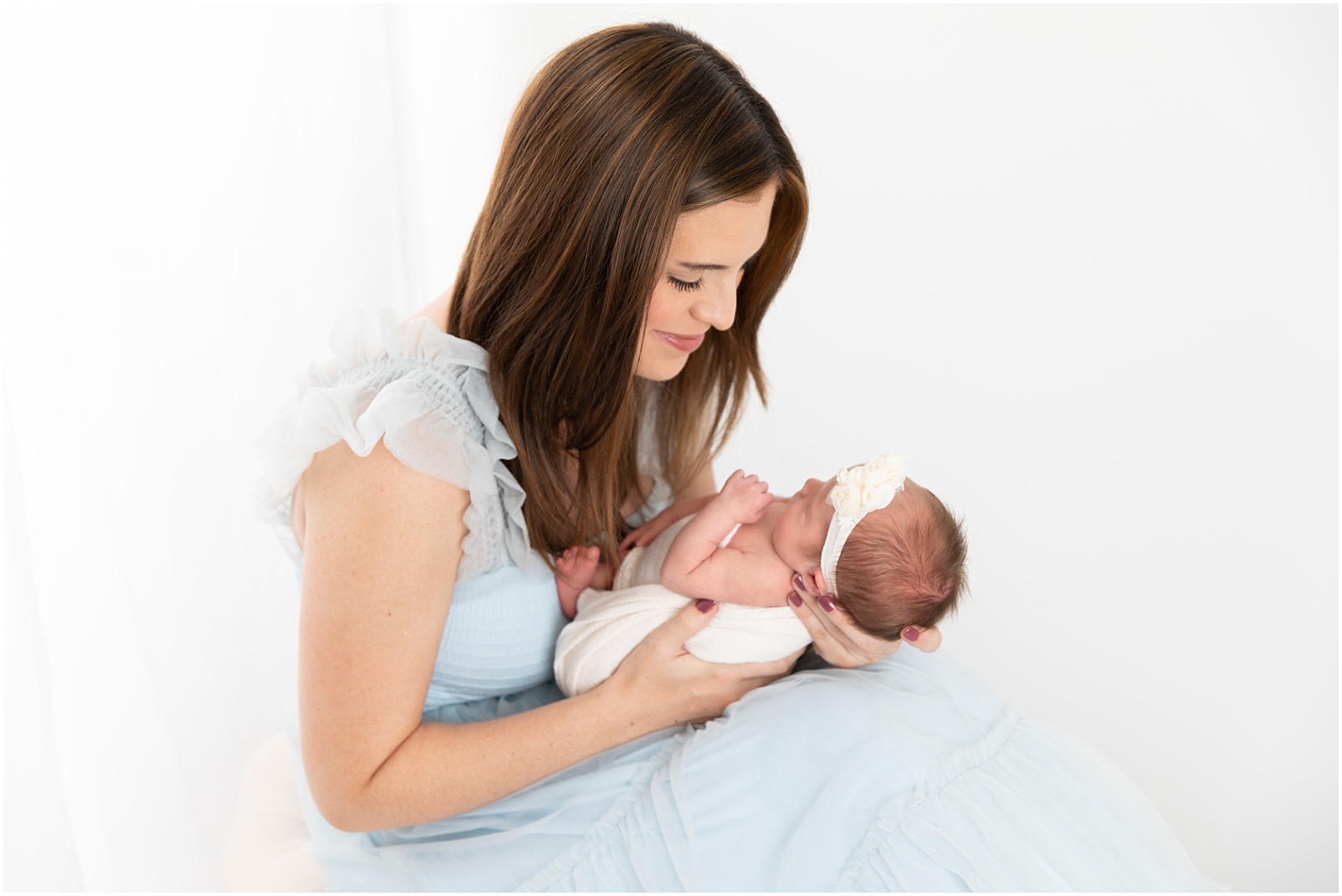 Mother wearing a light blue dress and holding her newborn girl swaddled in white with a floral headband