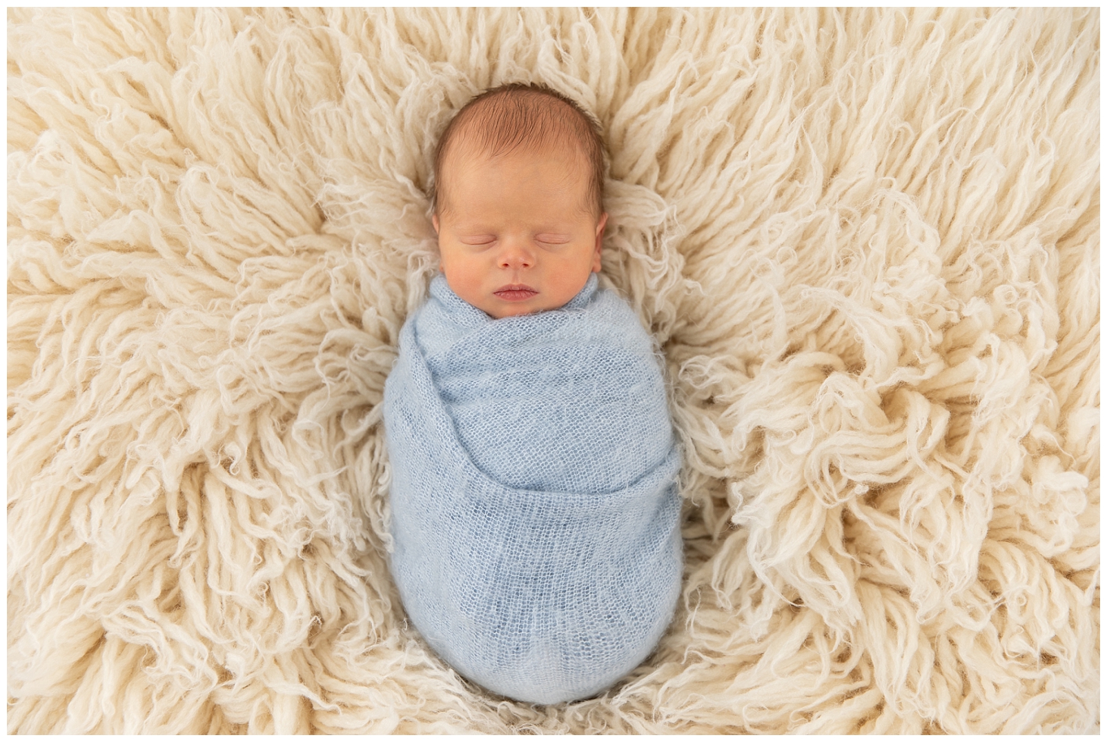 A newborn boy wrapped in a light blue knit fabric on a flokati