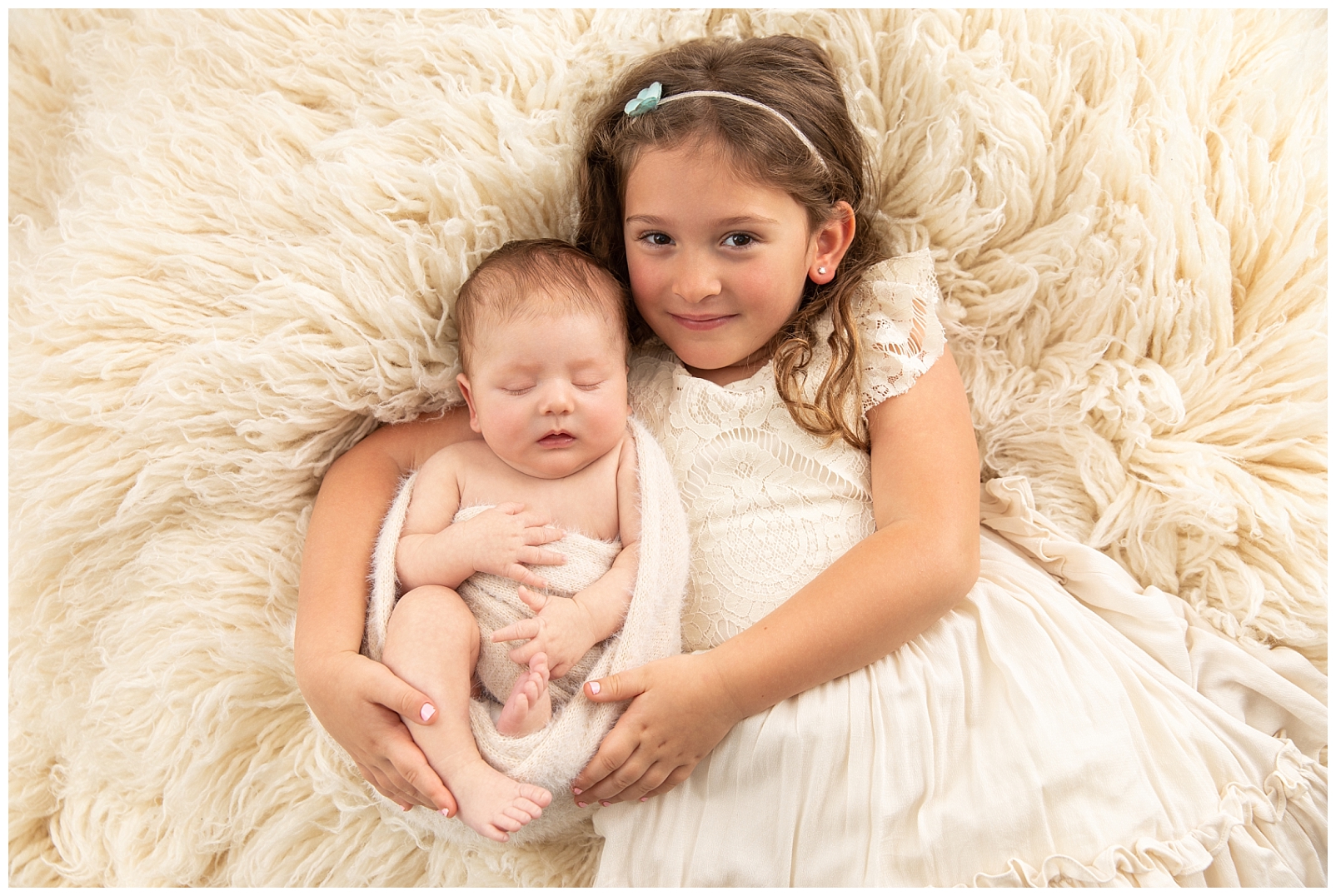 A girl and in a textured design dress holding her baby brother wrapped in ivory fuzzy fabric on a flokati background